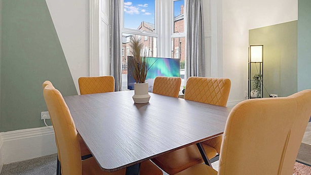 Un comedor con una mesa rectangular, cuatro sillas, un jarrón con plantas secas y vistas a edificios a través de amplios ventanales.