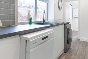 A kitchen with a dishwasher, a window with a view of a brick building, and a clock on the wall. - Charming 5-Bed Home in Sunderland by Stay with Us (Tyne and Wear)