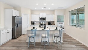 A modern kitchen with white cabinetry, a stainless steel refrigerator, a central island with bar stools, and a window with blinds.