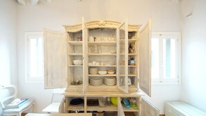 Une pièce avec un meuble blanc contenant des étagères remplies d’assiettes, de verres et d’ustensiles de cuisine. On aperçoit une chaise et une table au premier plan.