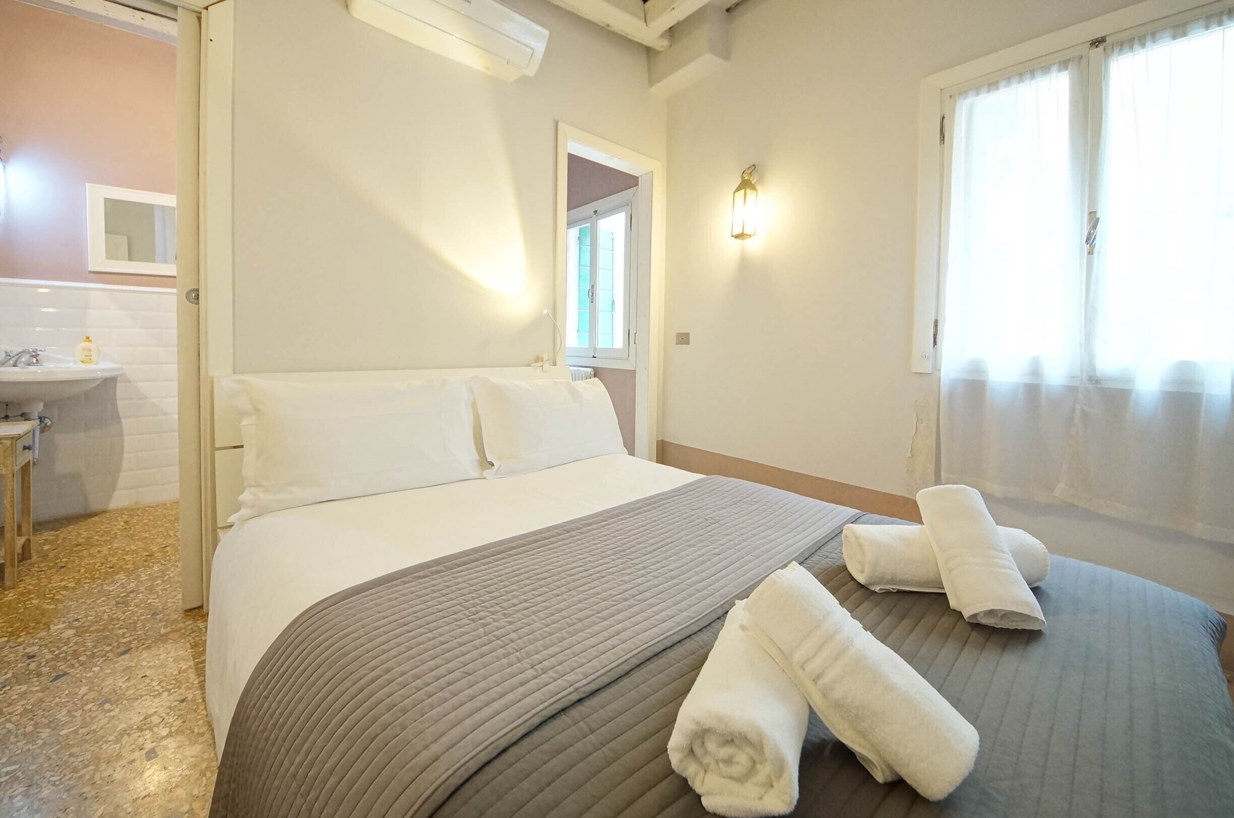 Una habitación de hotel con cama, mesita de noche, ventana con cortinas y baño con lavabo y espejo.