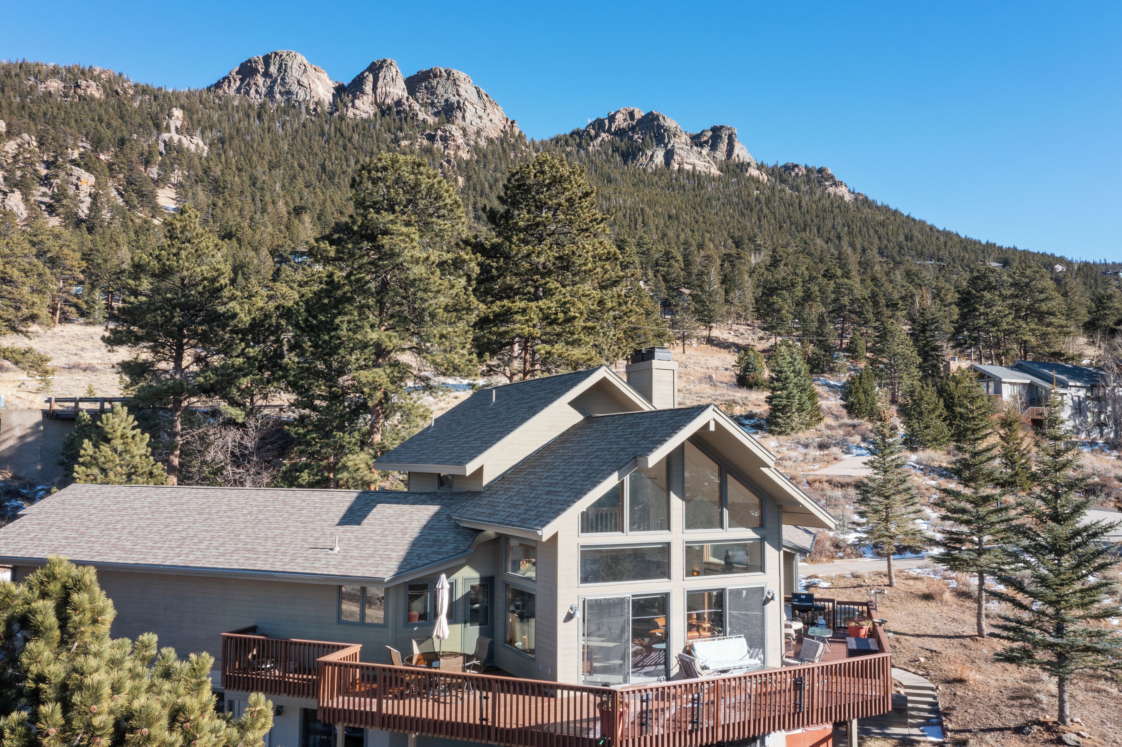 Une maison moderne à deux étages, dotée d’une grande terrasse et entourée de montagnes et de pins.