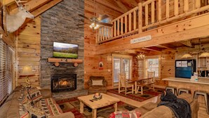 A cozy log cabin interior with a stone fireplace, a TV mounted on a wooden beam, a dining area, and a balcony with a sign reading "Hot Chocolate". - Scenic Hideaway: Pet-Friendly, Secluded, Game Room, fire pit, hot tub, Wi-Fi (Sevierville)