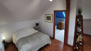A bedroom with a bed, a nightstand, a lamp, a painting, and a view of a bathroom through an open door. - Studio Spacieux, Route des Vins (Rosheim)