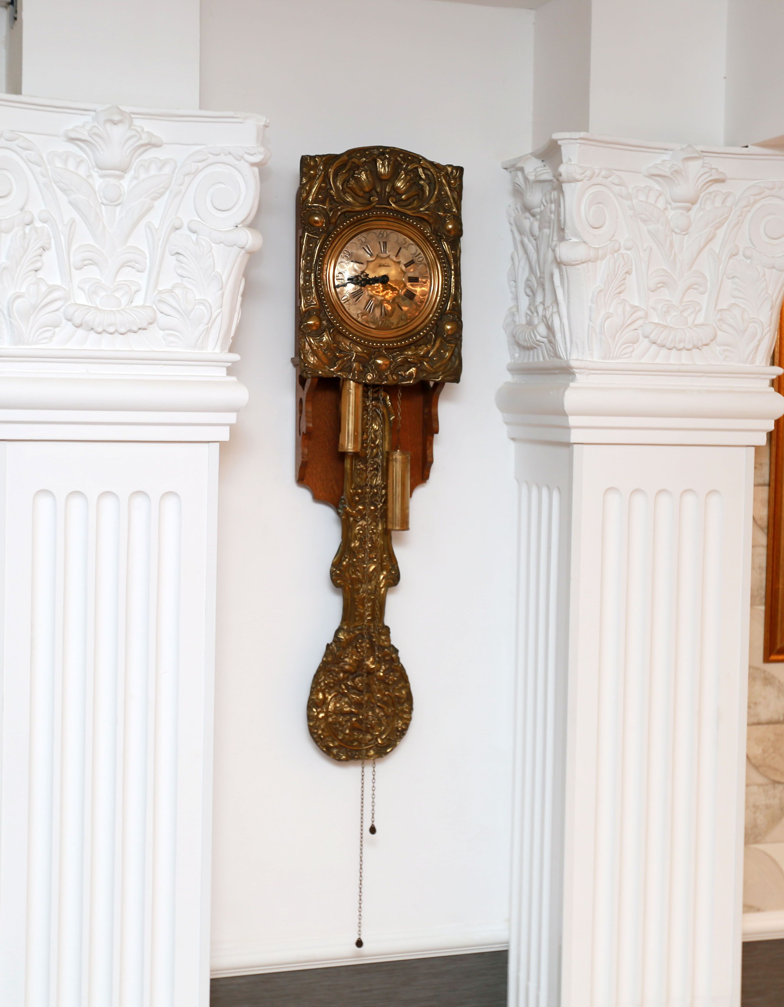 Une horloge ancienne fixée au mur, dotée d’un pendule, et placée entre deux colonnes blanches richement décorées.