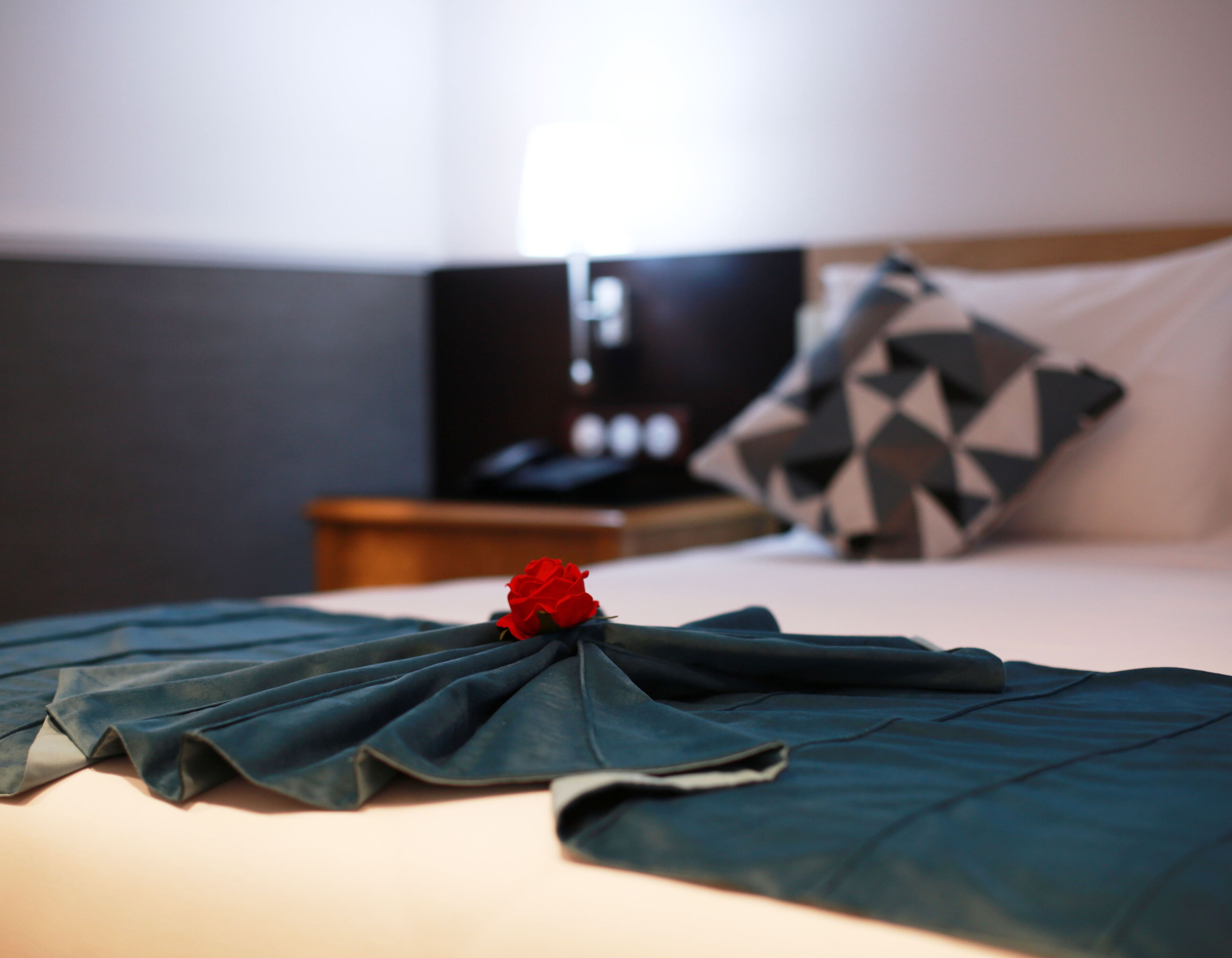 A neatly made bed with a dark blue bedspread, a red rose, and a geometric patterned pillow.