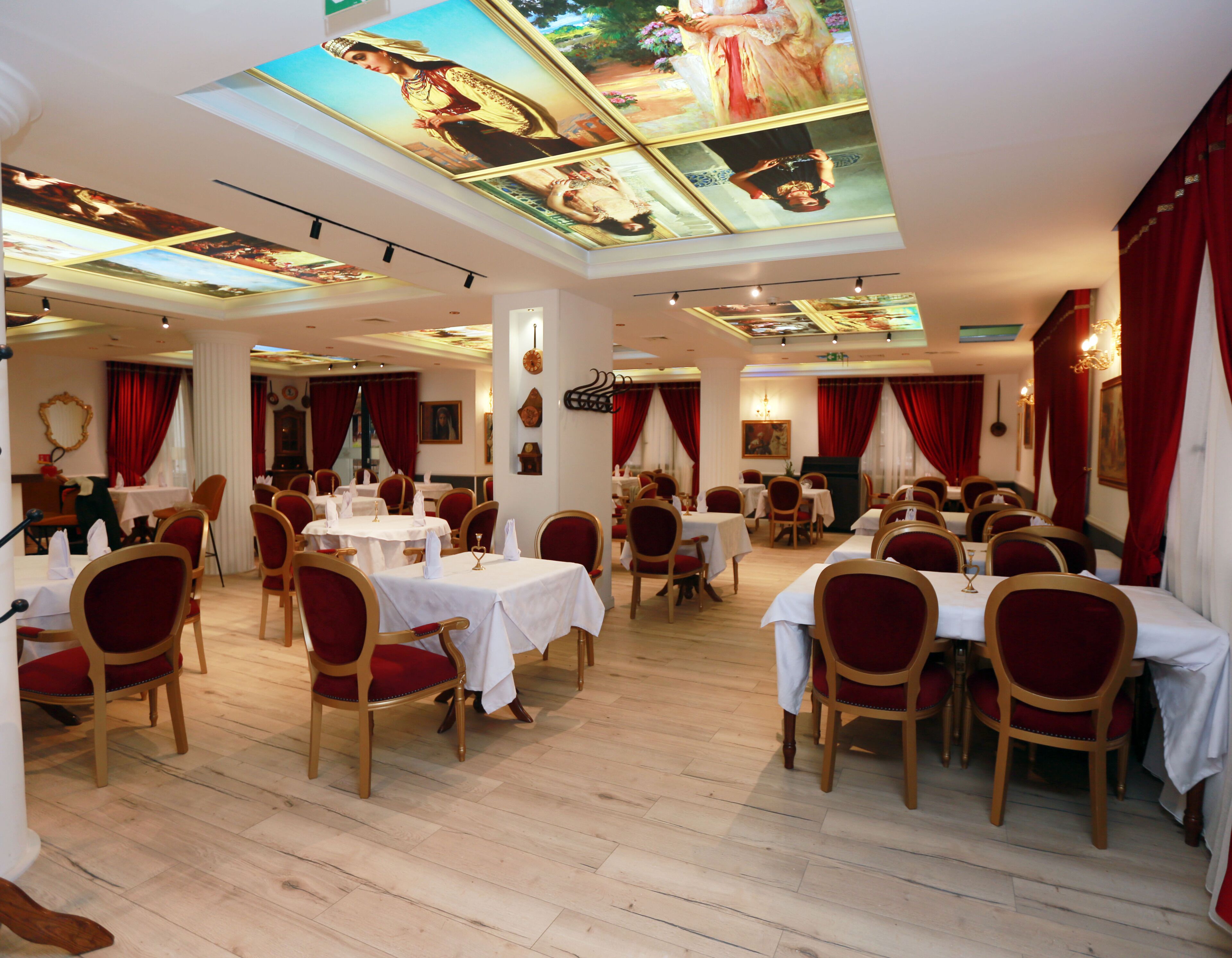 A restaurant with multiple tables and chairs, a high ceiling with artwork, and red curtains.