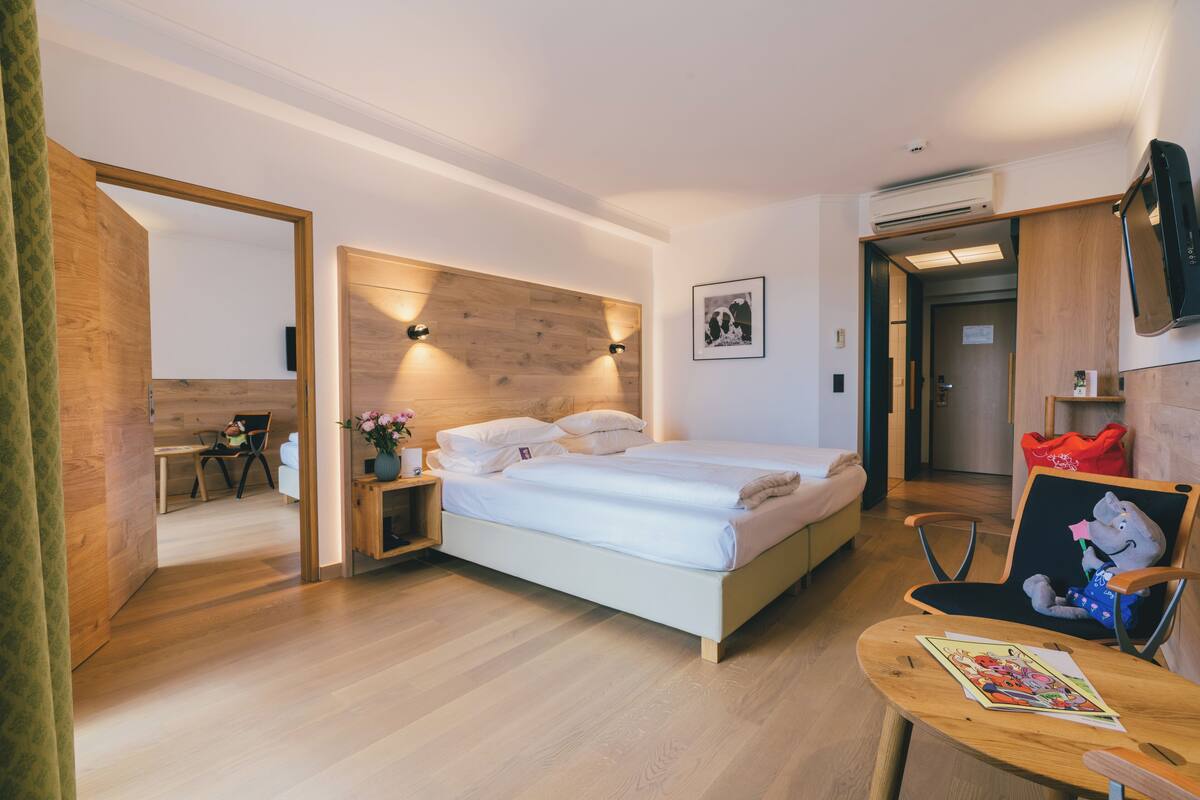 Een moderne hotelkamer met een houten hoofdeinde, een bed met witte lakens, een klein houten tafeltje met een boek, een stoel met een knuffel, en een aan de muur bevestigde televisie.