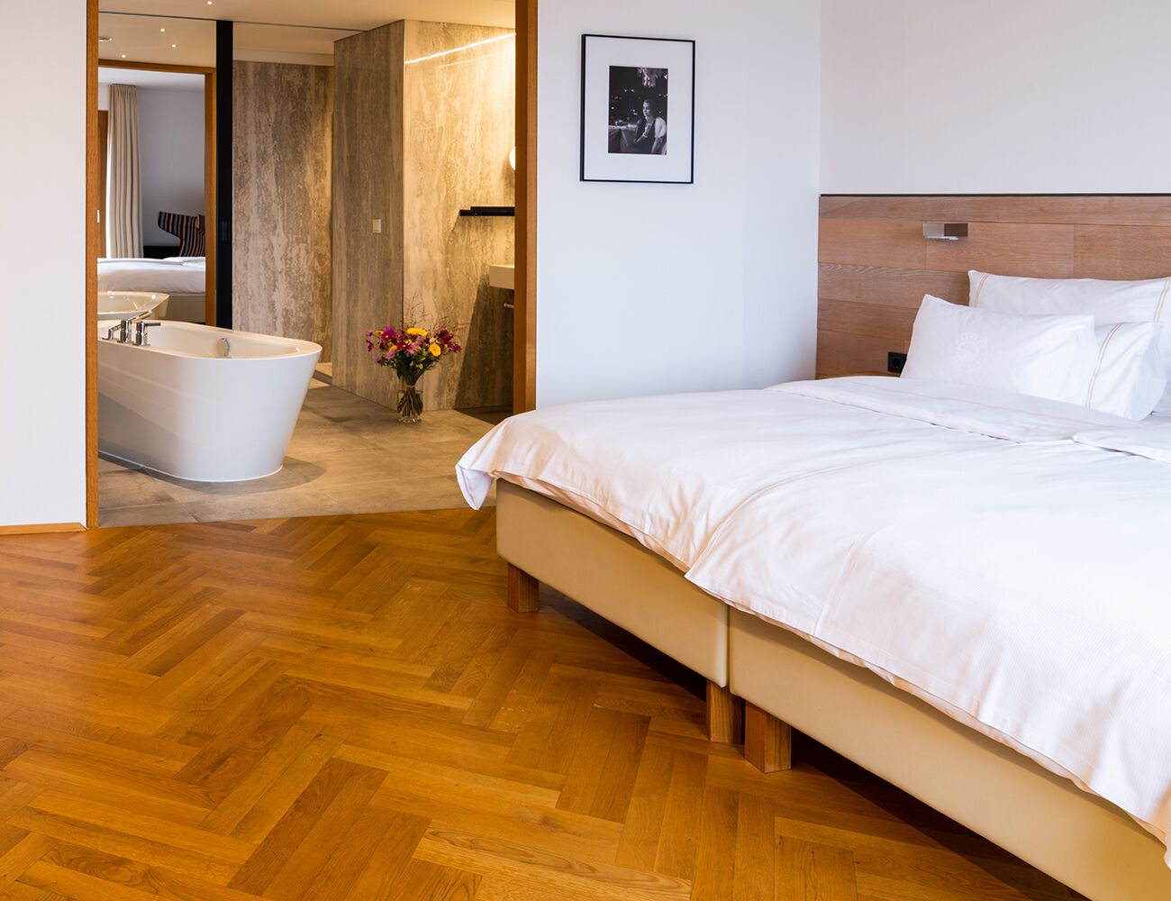 Una habitación de hotel moderna con suelo de madera, una cama grande con ropa blanca, cabecero de madera, bañera blanca y un cuadro enmarcado en la pared.