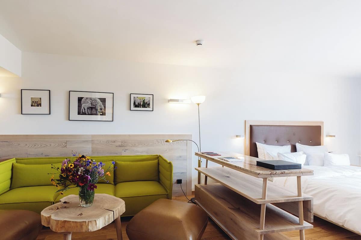Een moderne hotelkamer met een groene bank, een houten bijzettafeltje met een bloevas, een houten bureau met een laptop en een bed met witte lakens.