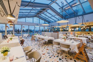 Un restaurante con mesas preparadas para comer, ventanas amplias y un techo alto con luces colgantes.