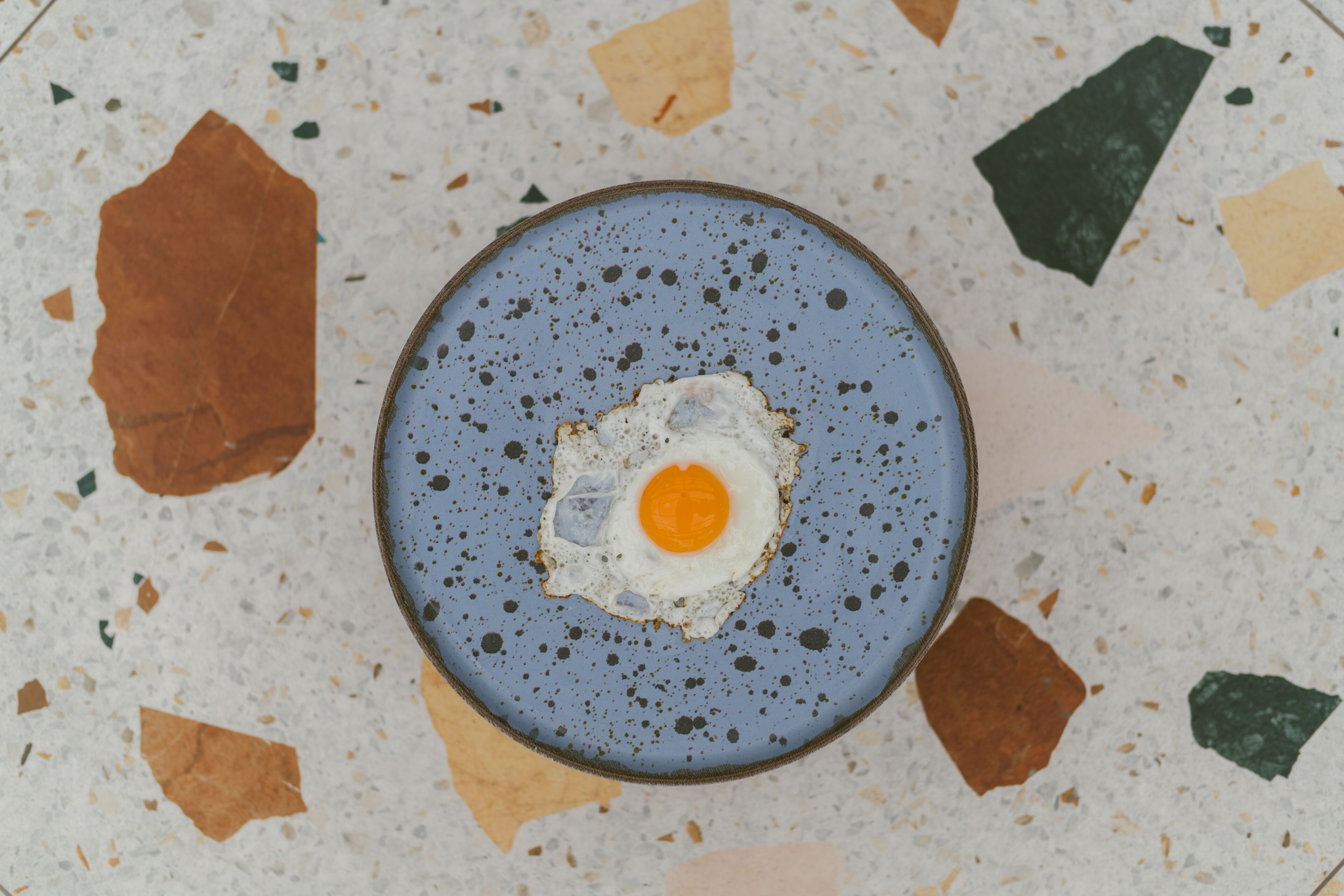 A blue plate with a fried egg on a speckled terrazzo floor.