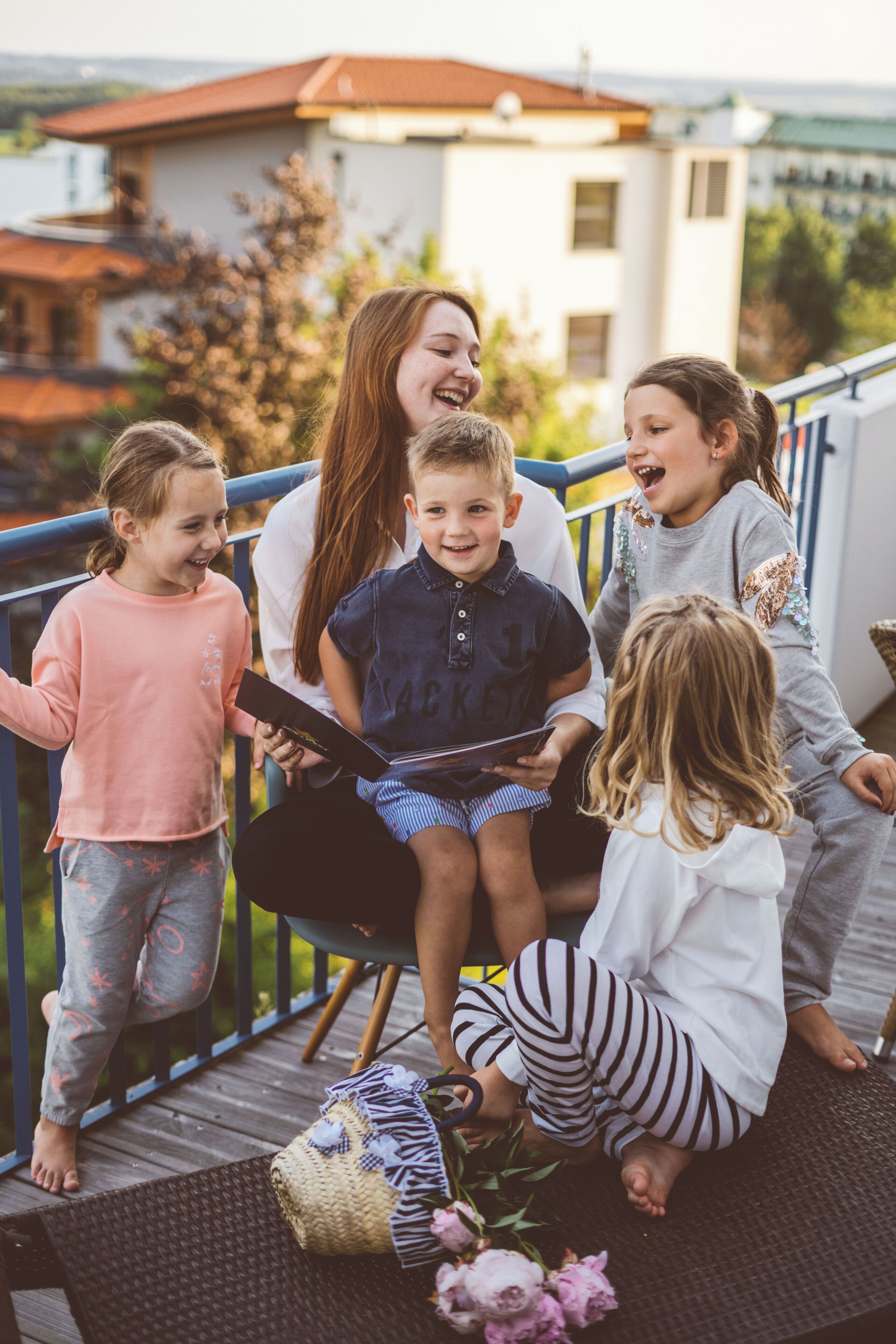 Een groep kinderen zit op een houten terras met een rieten stoel en een mand vol bloemen.