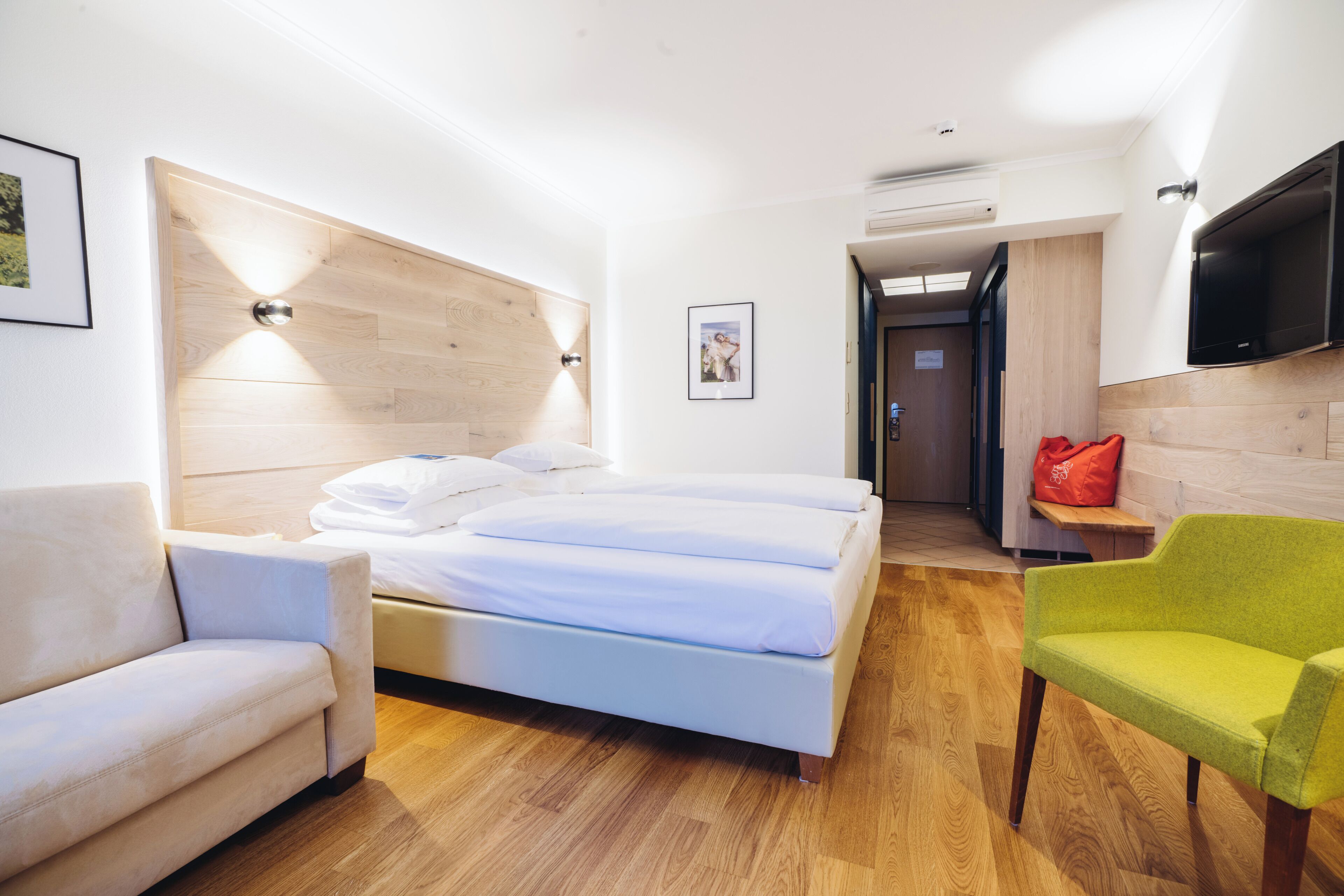 A modern hotel room with a wooden headboard, a bed with white linens, a green armchair, and a flat-screen TV mounted on the wall.
