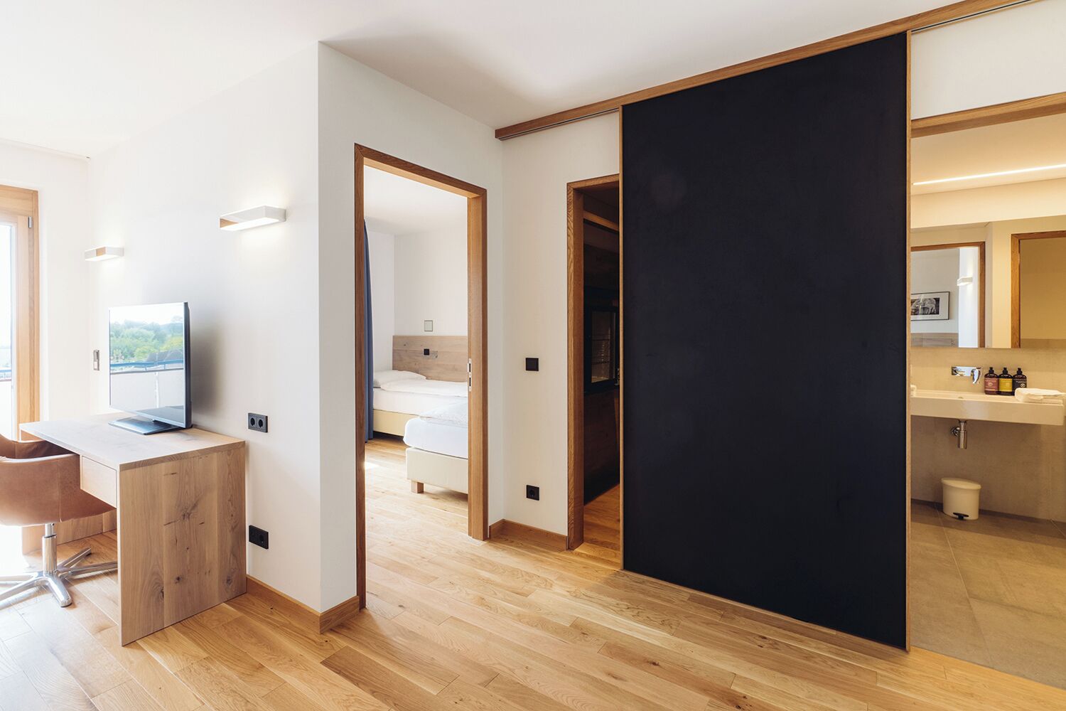 Una habitación de hotel moderna con un escritorio de madera, una cama y un baño con un espejo y lavamanos.