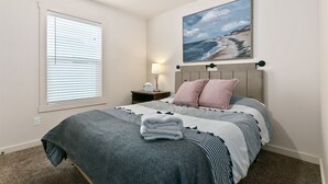 A bedroom with a bed, a nightstand, a lamp, and a painting of a beach scene. - 3 Adjacent Homes | Great for Families | Sleeps 24! (Meridian)