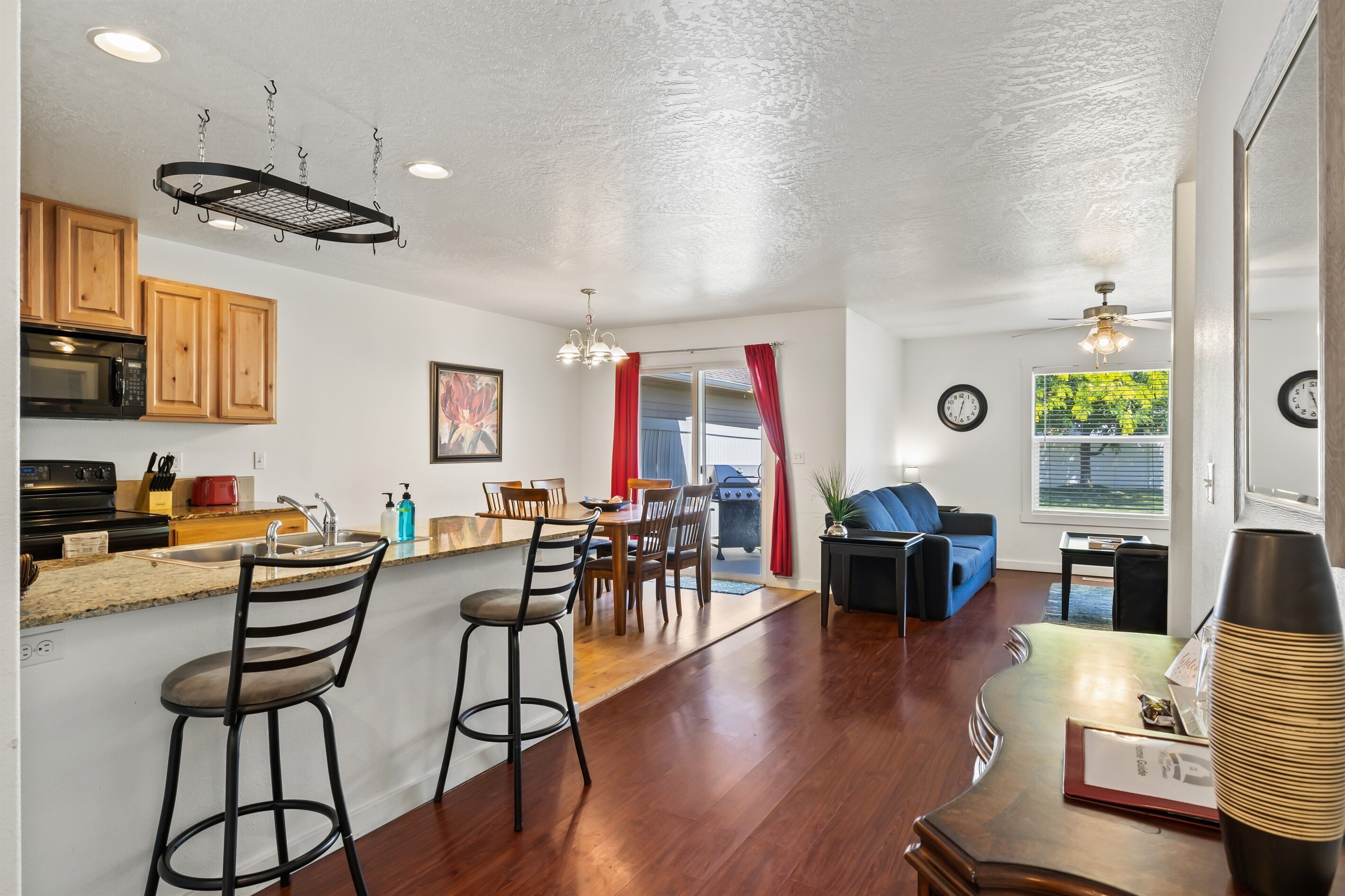 A modern kitchen with wooden cabinets, a microwave, and a hanging rack. There is a dining area with a table and chairs, and a living room with a blue couch and a coffee table.