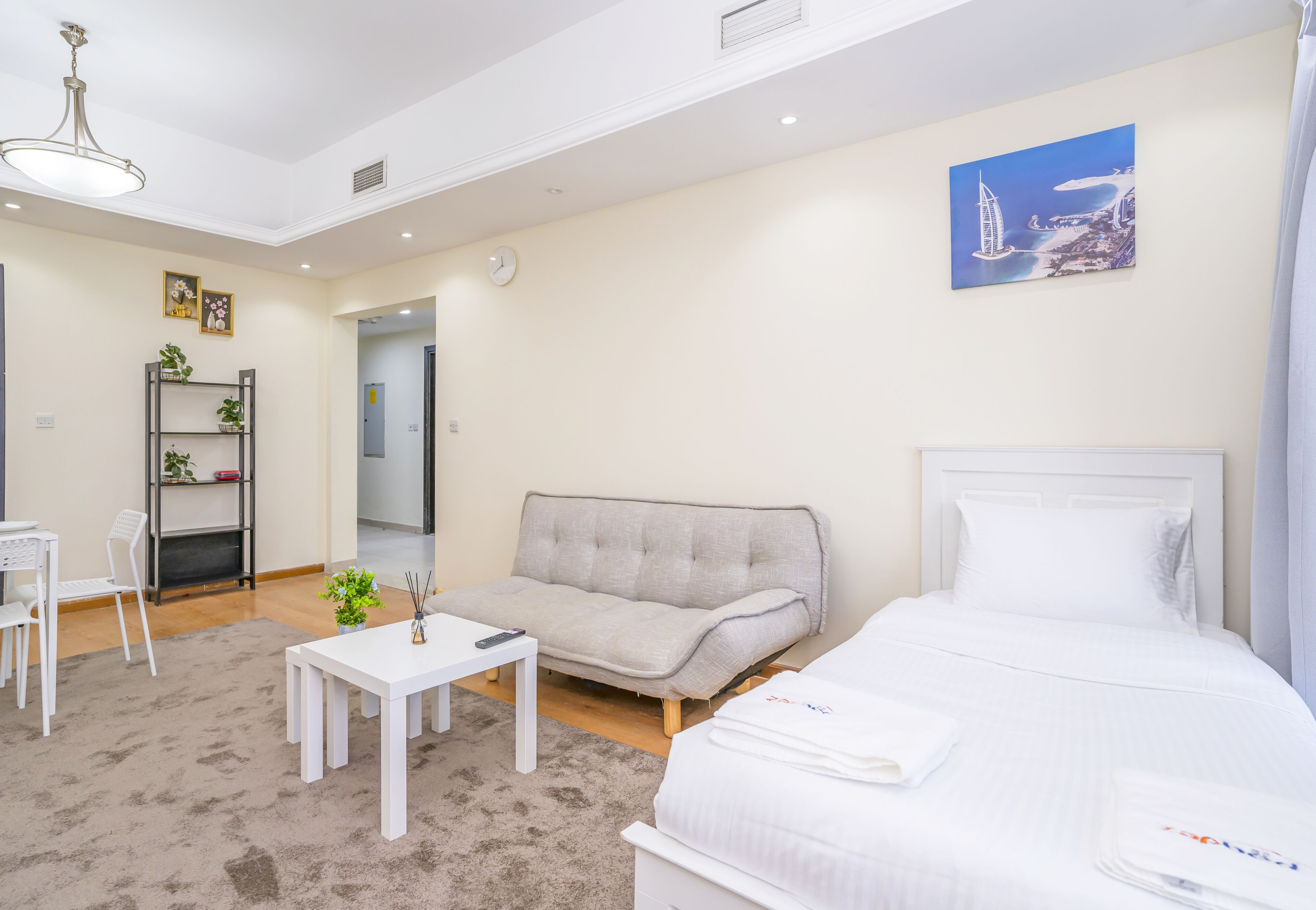 Habitación de hotel con cama, sofá, pequeña mesa, silla, escritorio, estante con plantas y un cuadro en la pared.