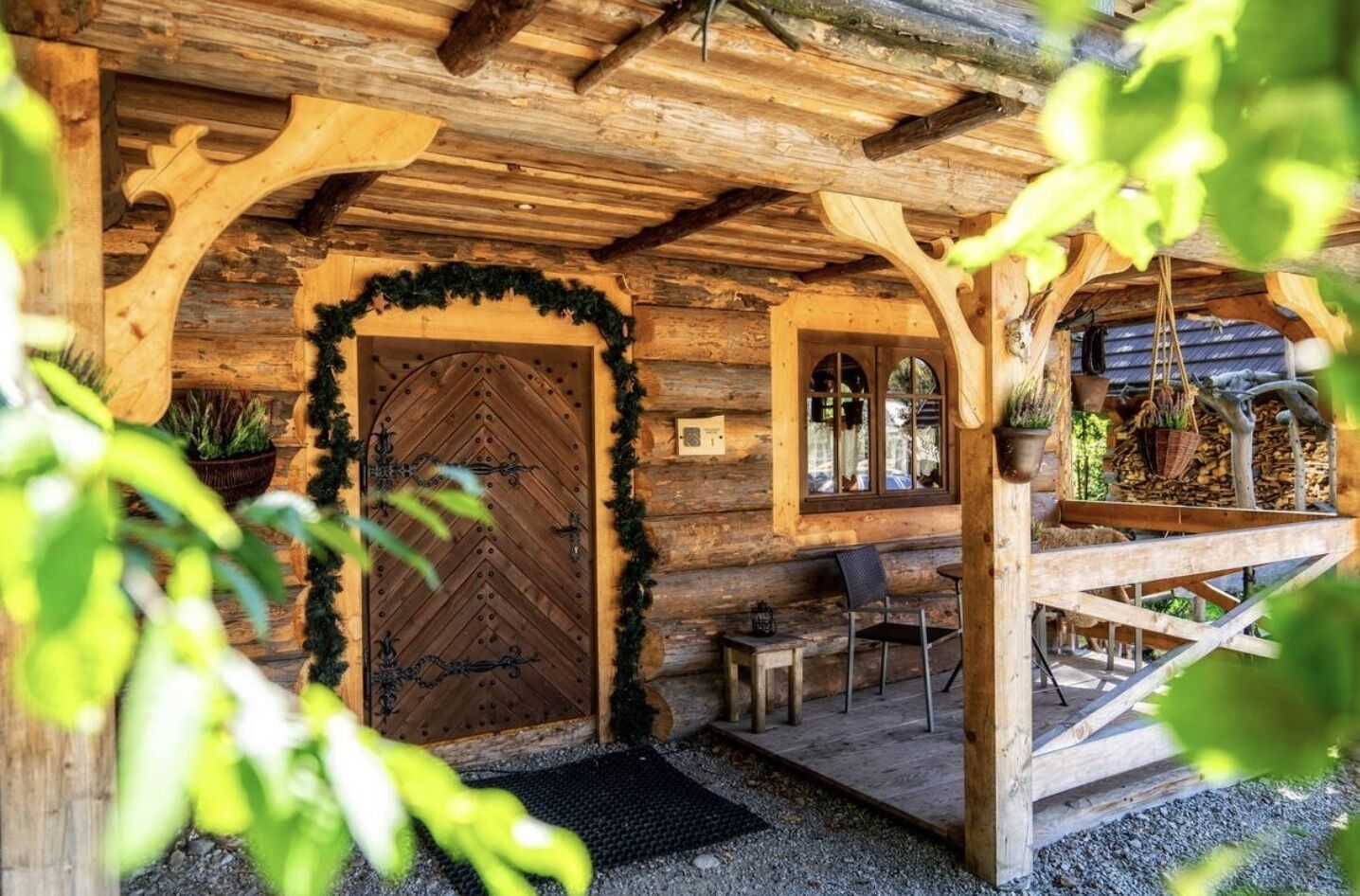 A rustic wooden cabin with a large wooden door, a small porch with a bench and table, and a window overlooking the cabin.