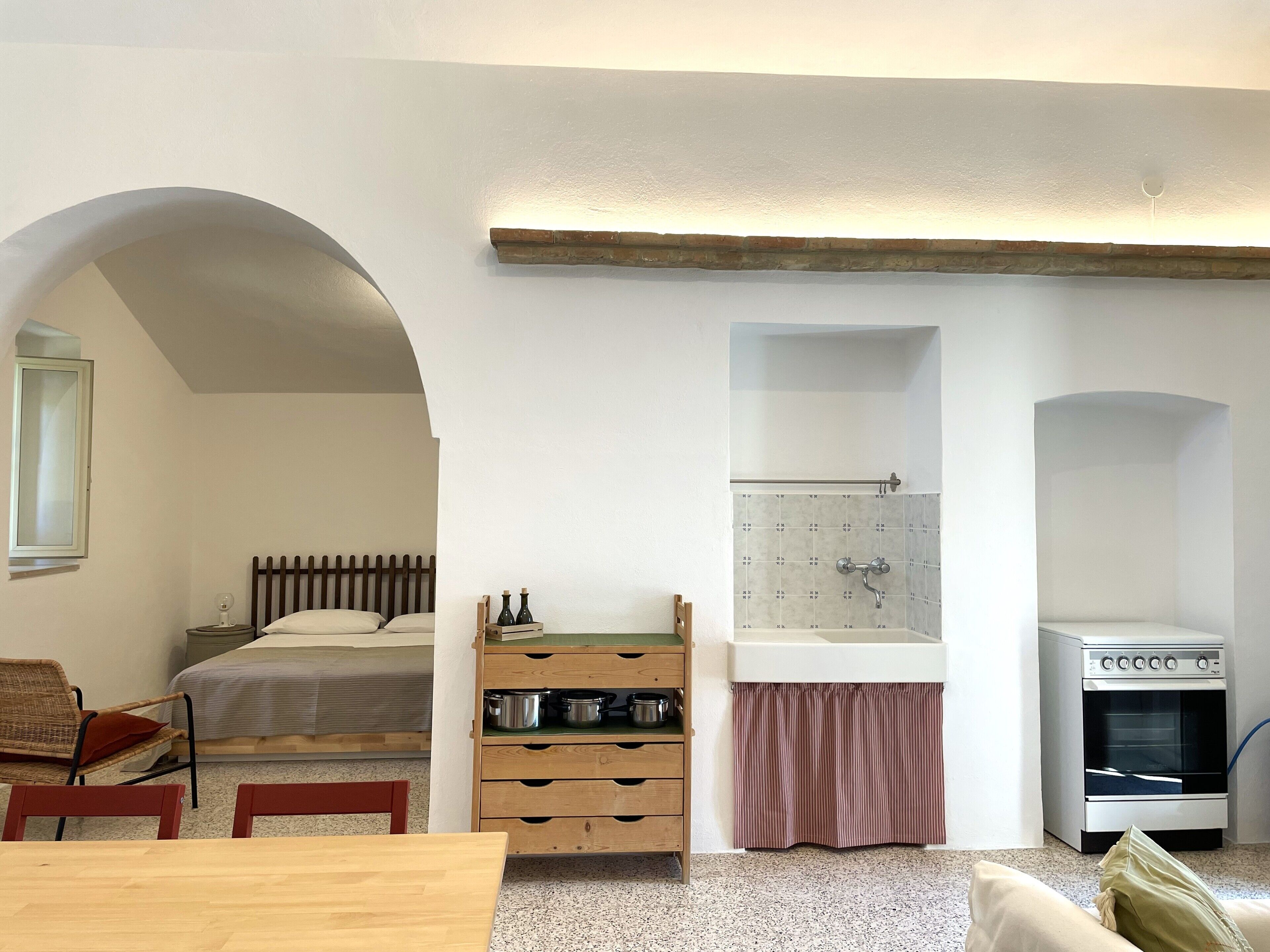 Een kleine kamer met een bed, een houten tafel, een kitchenette met een oven, en een badkamer met douche en wastafel.