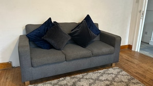 A grey sofa with dark cushions against a plain wall, a wooden floor, and a patterned rug. - Balham Double Deluxe Room 1 (London)