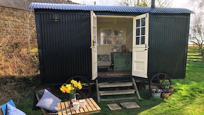 A small, open-fronted shed with a door leading to a kitchen area. There is a wooden table with two chairs and a vase of flowers outside. - Cosy Romantic Shepherd hut in Open Countryside (Newcastle-upon-Tyne)