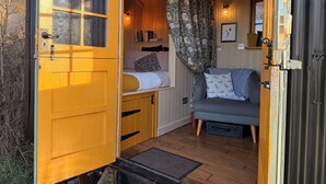 A cozy interior with a wooden door, a bed with a patterned pillow, a sofa with patterned cushions, and a wall-mounted shelf with books. - Cosy Romantic Shepherd hut in Open Countryside (Newcastle-upon-Tyne)