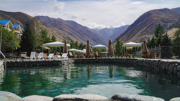A pool area with lounge chairs and umbrellas, surrounded by mountains and a clear sky. - Mountain View Hot Spring Resort (Koy-Tash)