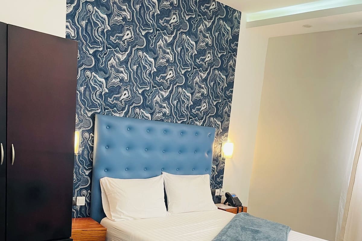 A neatly made bed with white linens and a blue headboard, a wooden nightstand with a phone, and a dark wardrobe.
