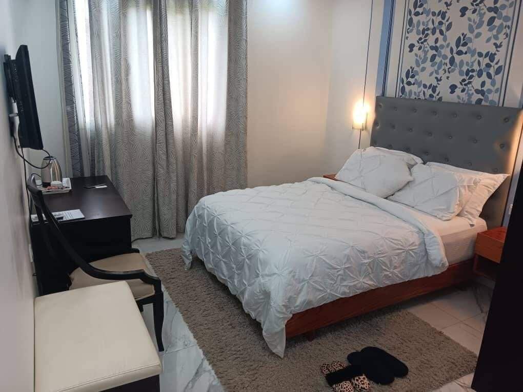 Ein Schlafzimmer mit einem Bett, einem Schreibtisch und einem Stuhl. Ein Fenster mit Vorhängen ist vorhanden.