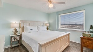 A bedroom with a bed, a nightstand, a lamp, a ceiling fan, and a window with a view. - Royal Palms 301 by Liquid Life (Gulf Shores)