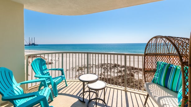 A balcony with turquoise chairs, a small table, and a wicker sofa with striped cushions, overlooking a beach and a pier. - Royal Palms 301 by Liquid Life (Gulf Shores)