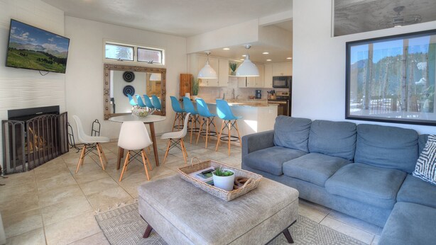 A modern living room with a grey sofa, a dining area with blue chairs, and a kitchen with white cabinets. - Grand Mountain Getaway | Ski, Fireplace, EV Charger (Durango)