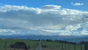 Paisaje rural con una carretera que se dirige hacia una cadena montañosa bajo un cielo nublado.