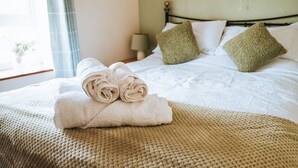 Una cama bien hecha con toallas enrolladas, un almohadón verde y un plaid beige sobre una manta texturizada.
