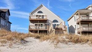 A beachfront house with a prominent balcony, a staircase leading to the entrance, and a sandy beach in the foreground.