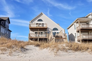 A beachfront house with a prominent balcony, a staircase leading to the entrance, and a sandy beach in the foreground.