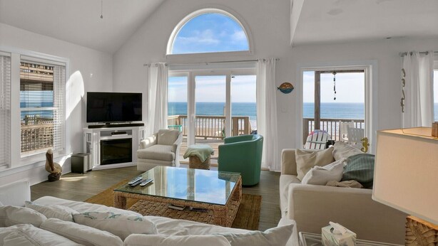 A spacious living room with a large TV, comfortable seating, and a view of the ocean through large windows.
