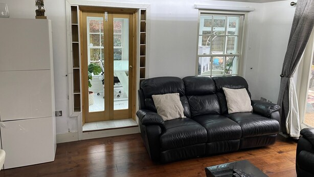 A living room with a black leather sofa, a coffee table, and a white refrigerator. There are two windows with curtains, and a door leading to a garage. - Lovely 2-bed Apartment in Brixton Central Location (London)