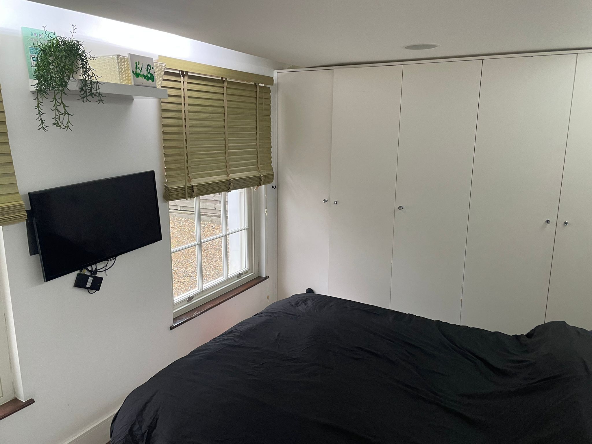 A bedroom with a bed, a TV mounted on the wall, a window with blinds, and a wardrobe.