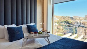 A modern hotel room with a large bed, dark headboard, and a view of a cityscape through a large window. - Penthouse Horizonte with stunning views in Colina del Sol (Cap Blanc Cullera)