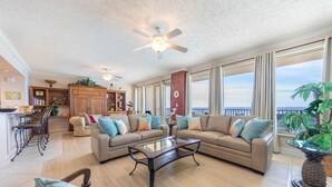 A spacious living room with a large sofa, a coffee table, and a ceiling fan. There is a kitchen area with wooden cabinets and a dining table with chairs. The room has large windows with a view of the ocean. - Hidden Dunes 11th Flr 3BR 3BA Slps 12!! (Panama City Beach)