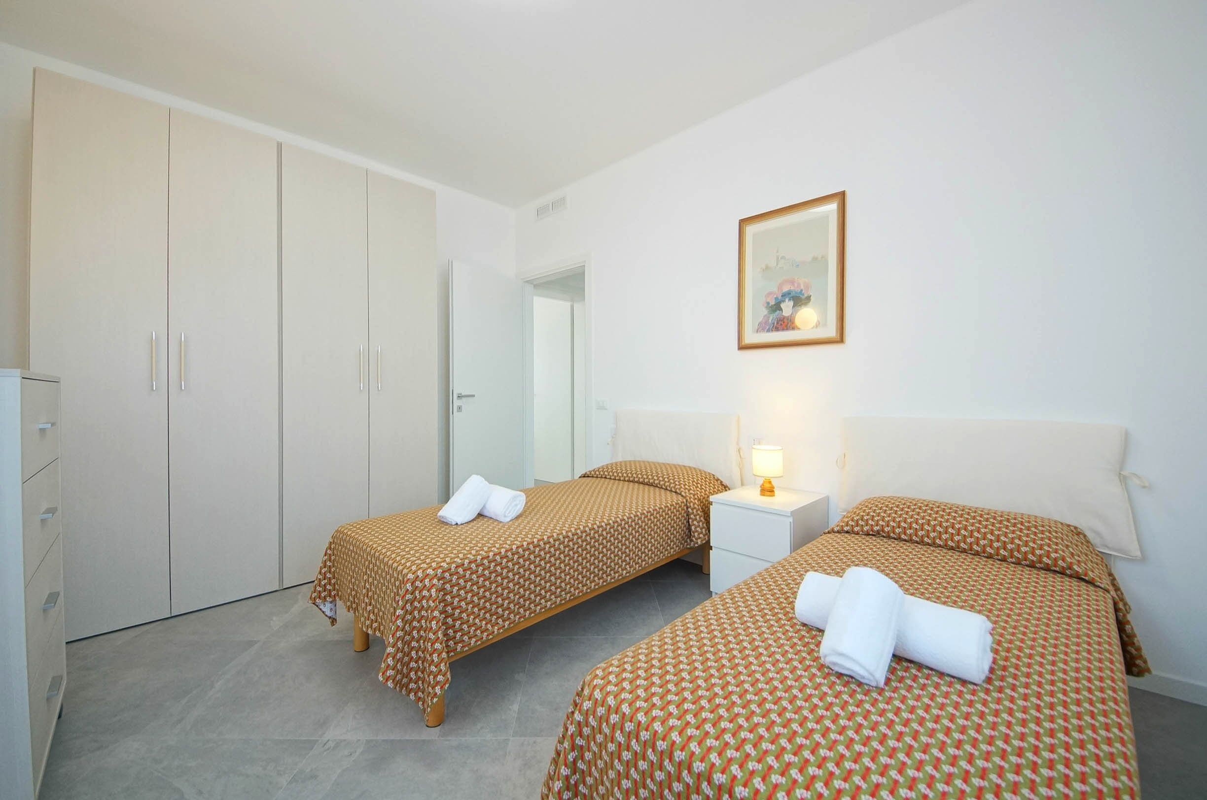 Habitación de hotel con dos camas, un armario, una mesita de noche con lámpara y un cuadro en la pared.
