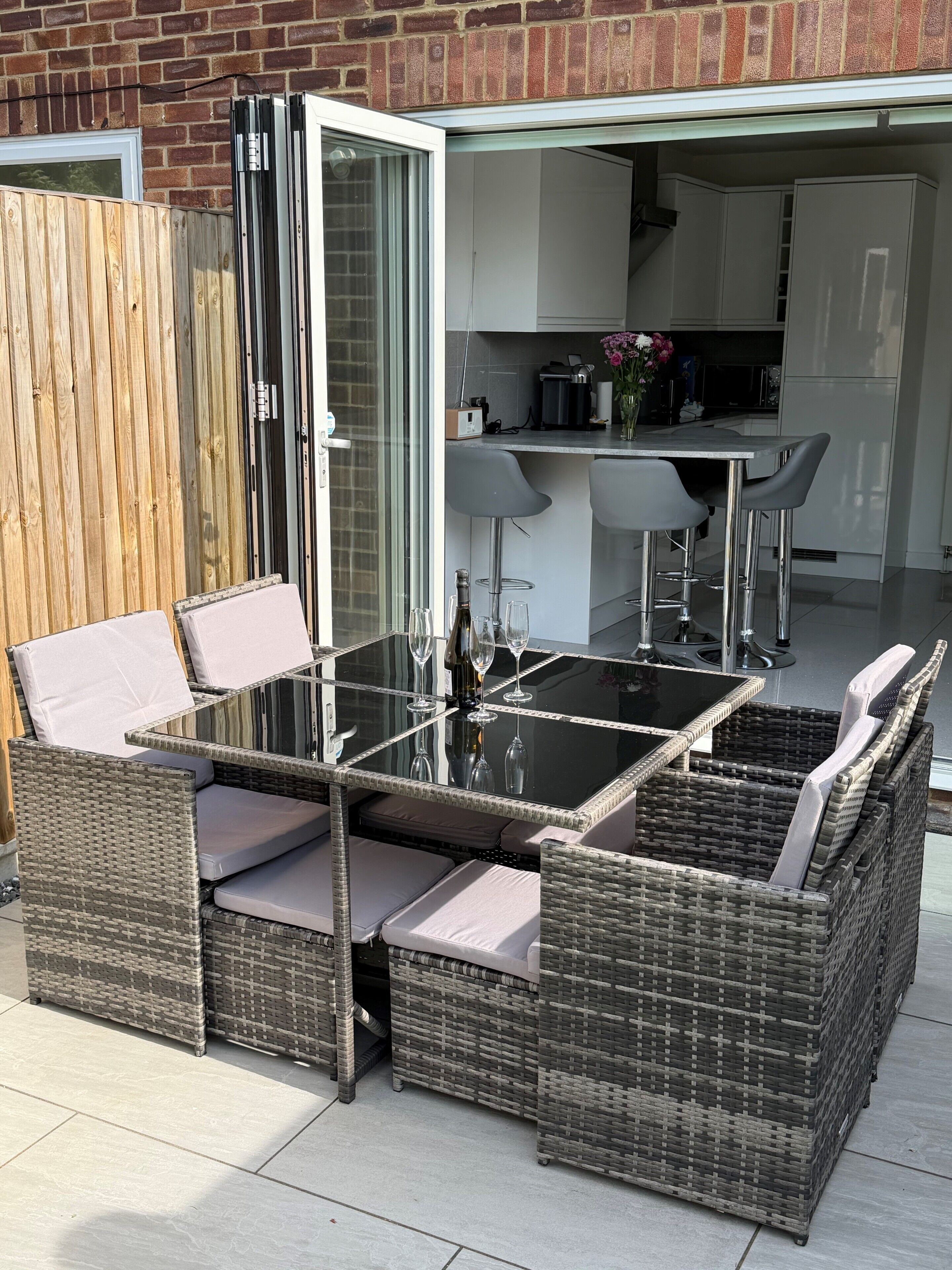 Un espacio al aire libre para comer con mobiliario de mimbre, una mesa de vidrio y vistas a una cocina y un garaje.