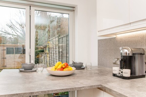 A modern kitchen with a large window, a coffee machine, and a bowl of fruit on the counter.
