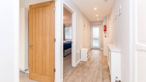 Un pasillo con puerta de madera, una cama con colcha azul, un radiador y un detector de incendios en la pared.