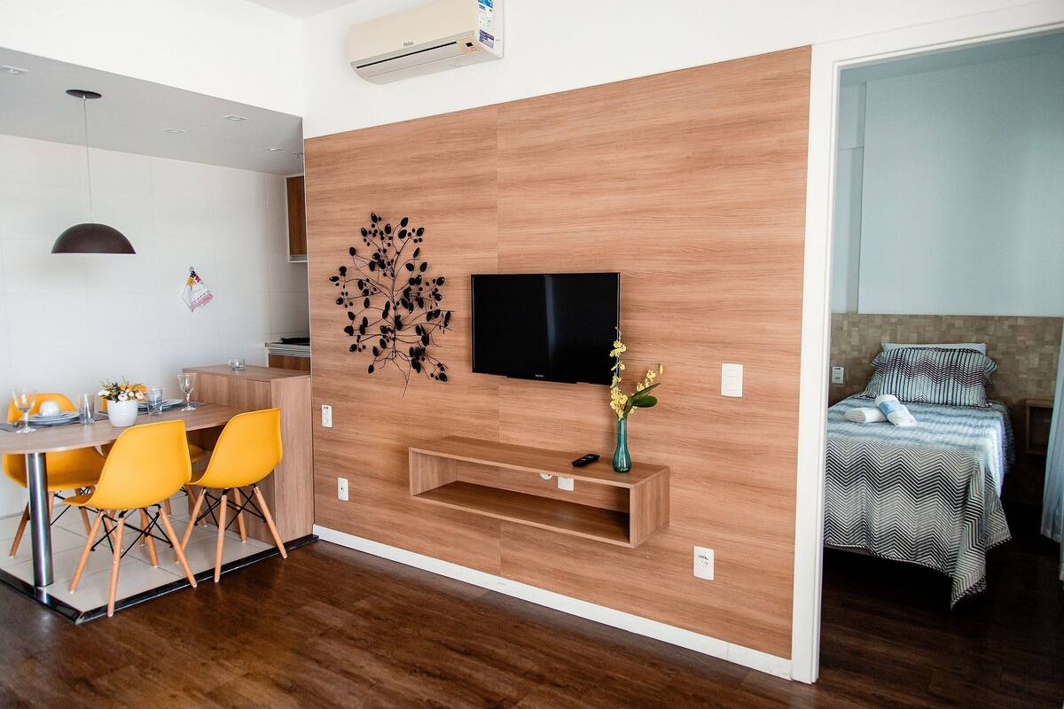 A modern living room with a wooden wall, a flat-screen TV, a dining area with yellow chairs, and a bed with striped bedding.