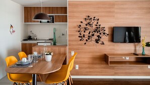 A modern kitchen with wooden cabinets, a dining table with yellow chairs, a wall-mounted TV, and a decorative plant. - Mondial Apartments By BnbHost (Salvador)