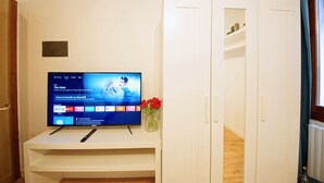 Ett rum med en tv på ett vitt tv-bord, en vit garderob och en vas med röda blommor.