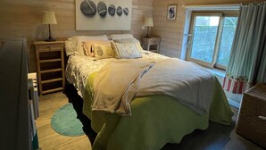 Un dormitorio acogedor con cama, dos mesitas de noche, lámpara, cabecera de madera, ventana con cortinas y una obra de arte colgada en la pared.